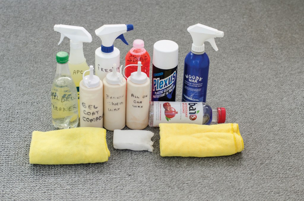 A high-angle, close-up shot of an assortment of boat detailing and cleaning supplies neatly arranged on a textured gray carpet. In the foreground, two folded yellow microfiber cloths sit on either side of a small white rectangular cleaning sponge. Behind them are several squeeze bottles and spray containers, many with handwritten labels such as "Mildew Cleaner," "Gel Coat Compound," "Banana Shield Wax," "All In One Wax," "APC," "Freshener," and "Soap." The collection also includes commercial products like a can of Plexus Plastic Cleaner, a blue spray bottle of Woody Wax, and a clear Bai5 antioxidant beverage bottle resting horizontally on its side.