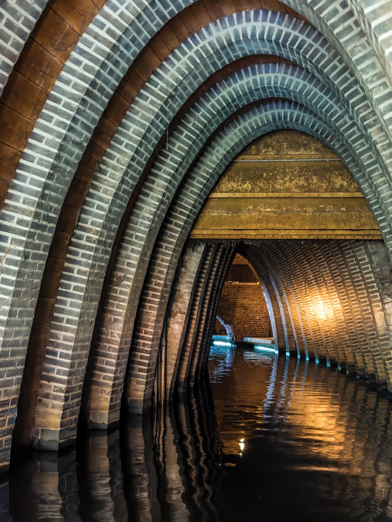 A dramatic vertical shot from water level looking through a subterranean canal vaulted by historic brick arches. The architecture features a series of closely spaced, pointed Gothic-style arches made of grey and brown bricks that curve over the dark, reflective water. Warm light illuminates the textured brick walls further down the tunnel, while a hint of cool, blue light glows at the water's edge in the background where the narrow waterway bends out of sight.