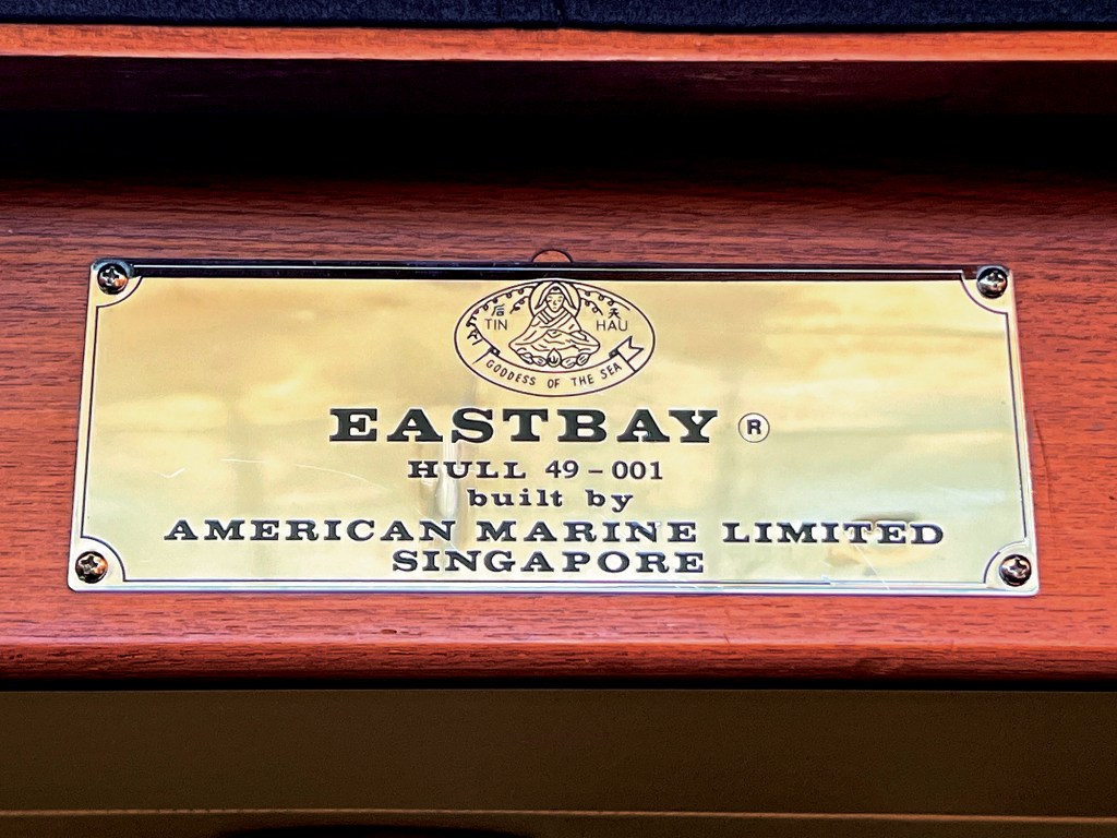 A close-up shot of a polished brass builder's plaque mounted with four screws onto a rich, reddish-brown wood surface. The plaque features an oval emblem at the top depicting "Tin Hau, Goddess of the Sea," followed by text engraved in bold black lettering that reads: "EASTBAY®, HULL 49 - 001, built by AMERICAN MARINE LIMITED, SINGAPORE".