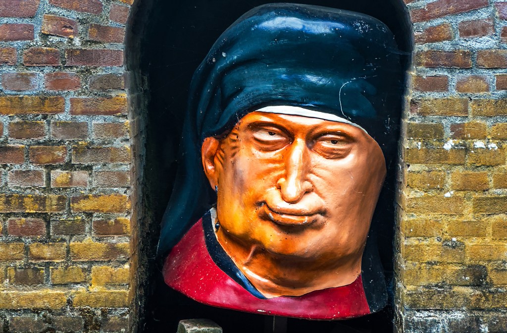 A close-up shot of a large, stylized sculptural bust of a person's head nestled inside a dark brick alcove. The sculpture depicted has an intense, slightly smirking facial expression with narrow eyes, a prominent nose, and warm orange-toned skin. The figure wears a draped black or dark blue headscarf or cowl and a red collar piece. The sculpture is framed tightly by an weathered, multi-toned brown and yellow brick wall.