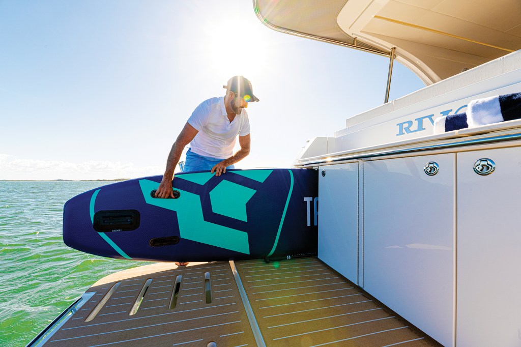A sunny, low-angle shot of a man wearing a white polo shirt, blue shorts, and a black baseball cap standing on the teak-planked swim platform of a yacht. He is sliding a large, dark blue and turquoise stand-up paddleboard into a dedicated, built-in horizontal storage locker located beneath the transom seating. The sun flares brightly overhead against a clear blue sky, illuminating the calm green water surrounding the boat.