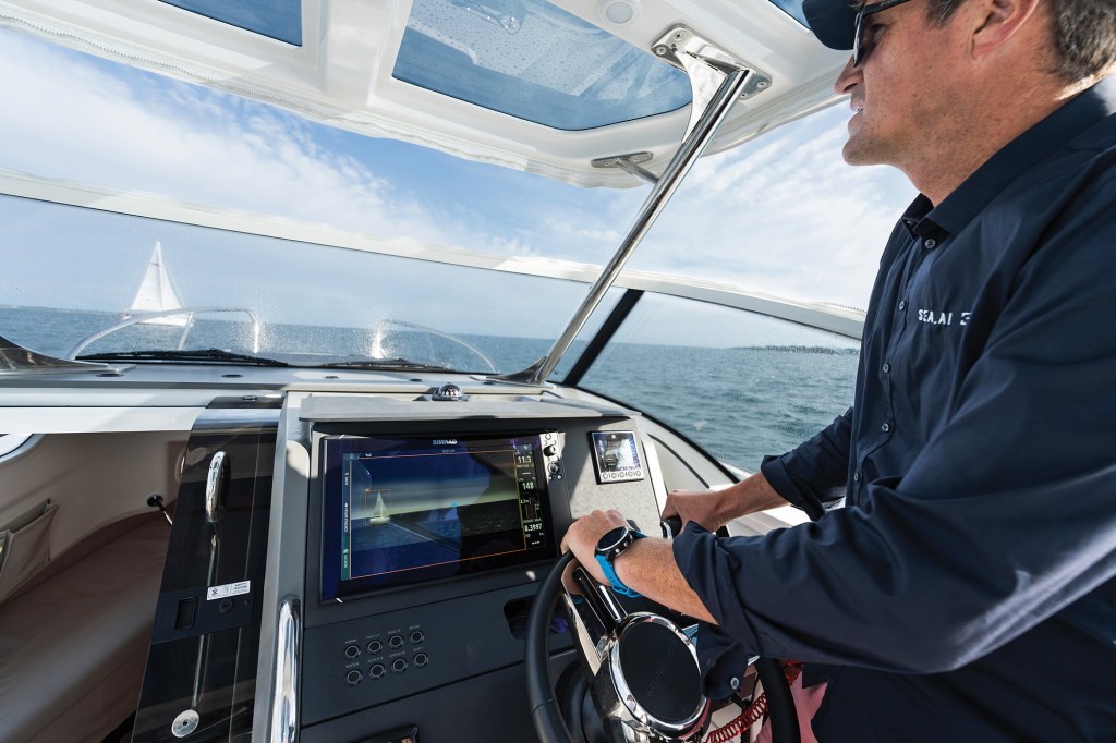 A close-up, side-profile view of a skipper steering a motorboat through open water from the helm station. The man wears a navy blue shirt with "SEA.AI" branding, sunglasses, and a blue sports watch as he grips the black steering wheel. The boat's dashboard features a prominent Simrad multifunction display screen showing an advanced thermal or AI-assisted navigation overlay that highlights a white sailboat on the water ahead. The same white sailboat is visible in the distance through the boat's clear front windshield under a partly cloudy sky.