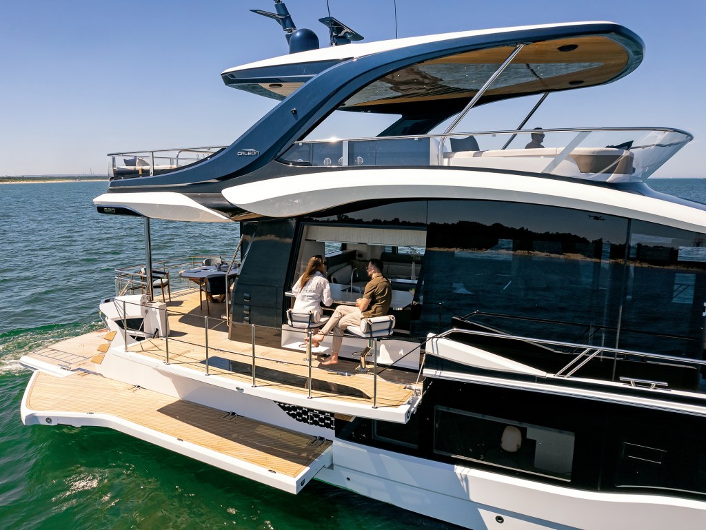 A high-angle, side-profile view of a luxury Galeon 620 Fly motor yacht anchored on calm green water under a clear blue sky. The vessel is in "beach mode," showcasing its innovative portside hull section dropped down horizontally to create an expanded teak-decked terrace balcony with silver safety railings. A man and a woman sit on modern bar stools at an open-air galley bar countertop that seamlessly blends the interior kitchen with the outdoor deck space. Overhead, the expansive flybridge structure features dark metallic blue accents and a wood-lined hardtop roof casting shade onto the main deck below.