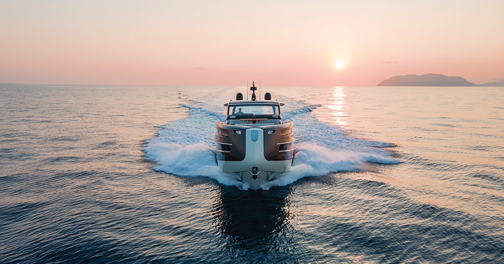 A wide, head-on shot from the water looking directly at the unique, inverted bow snout of an Invictus ST550 yacht cruising at speed. The dark-hulled luxury boat pushes up crisp white wake wings on both sides as it heads toward the camera. In the background, a warm sunset glows intensely over a calm, open ocean, casting a soft pink and orange hue across the sky and creating a glistening light path on the water's surface next to a distant mountain silhouette.