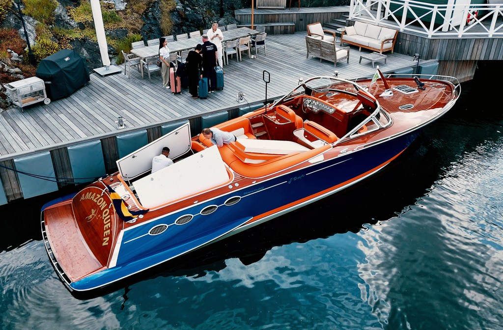 An elevated, high-angle view of the luxury motor yacht "Amazon Queen" docked alongside a wooden waterfront deck. The vessel features a striking Prussian blue hull, varnished mahogany woodwork, bright orange upholstery, and open engine hatches where crew members are working. On the adjacent dock, a small group of people stands with luggage near outdoor dining and lounge furniture.