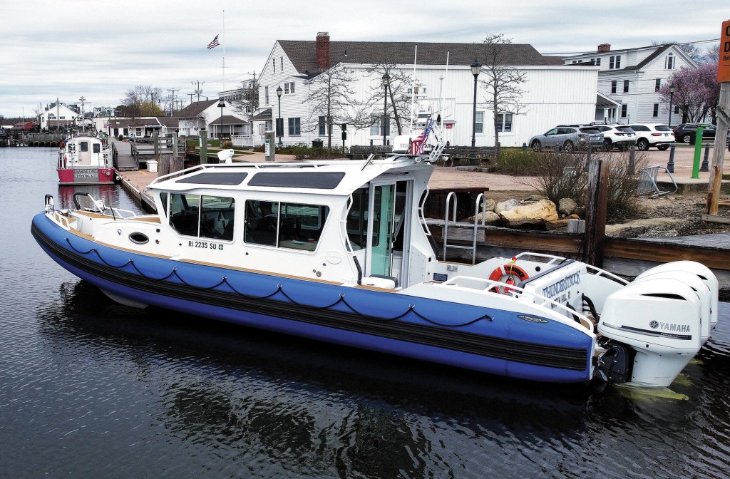 A three-quarter view of a Life Proof rigid inflatable boat (RIB) docked alongside a wooden pier. The vessel features a white enclosed cabin cruiser superstructure built onto a bright blue inflatable collar hull, powered by triple white Yamaha outboard motors at the stern. The transom displays the name "THUNDERSTRUCK." In the background, calm coastal waters are flanked by white coastal village buildings, parked cars, and an American flag flying at half-mast under an overcast sky.