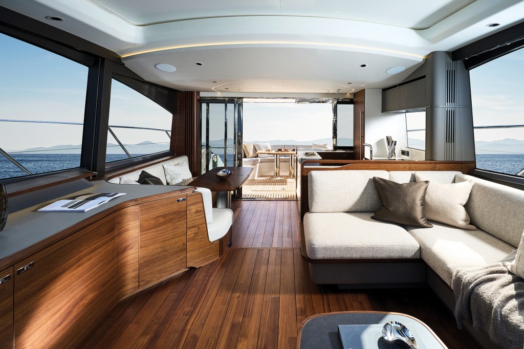 Modern main deck saloon of a Princess V65 yacht featuring rich hardwood flooring, a contemporary L-shaped sofa with neutral cushions, fine wooden cabinetry, and large panoramic windows looking out to the sea.