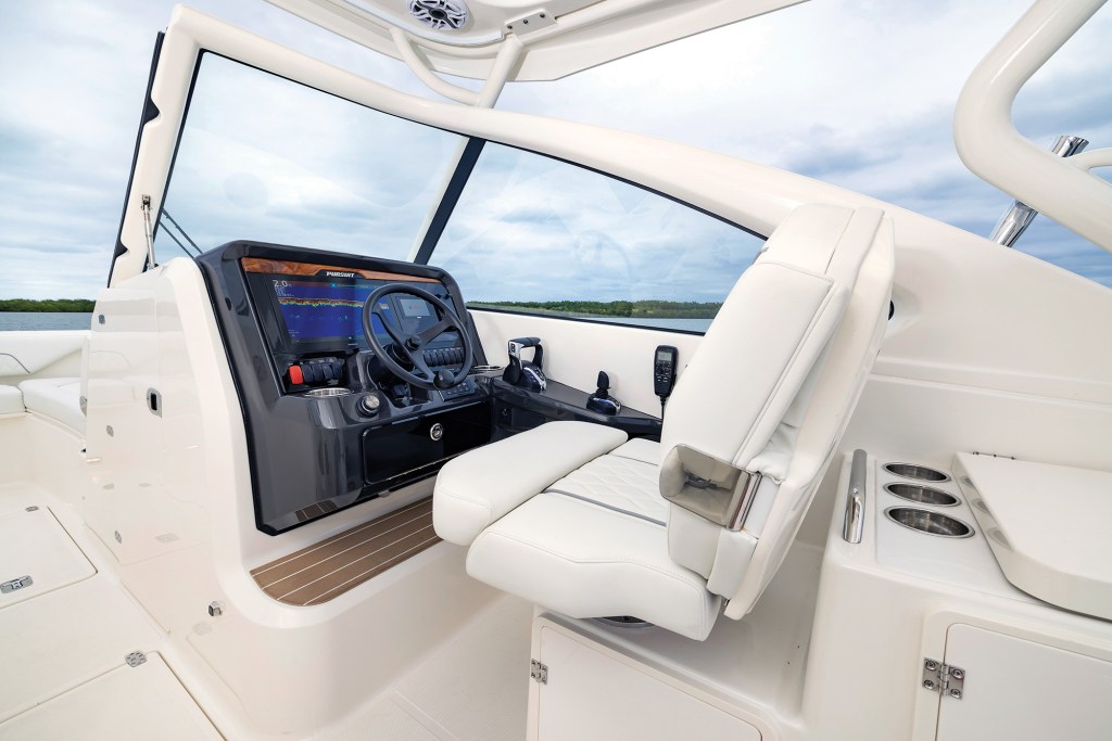 A wide-angle interior shot of the modern, clean helm station on a white Pursuit DC 286 dual-console boat. On the right sits a premium white captain's chair with diamond-quilted stitching and a folded-up bolster cushion. The dark grey dashboard features a black steering wheel, a large integrated digital navigation display showing active sonar data, a row of red and black rocker switches, electronic throttle controls, and a joystick docking controller. A large, high glass windshield wraps around the cockpit, offering a clear view of calm green water and a low-lying distant shoreline under a cloudy sky.