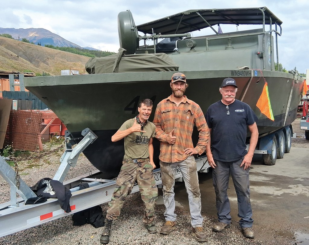 Three smiling men stand side-by-side posing in front of a large military Patrol Boat River (PBR) resting on a multi-axle boat trailer in a gravel yard. The military craft is painted in a matte olive drab green color with the black hull number "44" stenciled near the bow. The man on the left wears camo pants and an olive green t-shirt while flashing a "shaka" hand sign. The center man wears a brown plaid flannel shirt and gives a thumbs-up, and the older man on the right wears a black graphic t-shirt and baseball cap. In the background, grassy rolling hills rise up under a cloudy sky.
