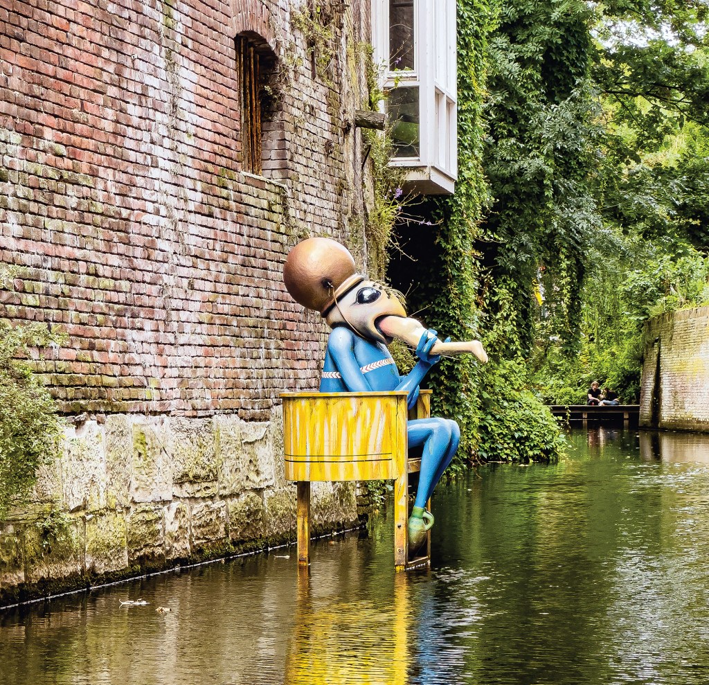 An outdoor, water-level view of a narrow canal canal lined with historic buildings and lush greenery. Standing directly out of the calm water on wooden stilts is a surreal, colorful sculpture of a blue creature with a bulbous, copper-colored head, sitting on a weathered yellow chair. The character appears to be swallowing a pale leg, referencing artwork by Hieronymus Bosch. To the left, a tall weathered red brick wall drops into the water, while the right bank is heavily covered in thick green climbing ivy and overhanging trees. In the background, a small wooden dock spans across the quiet waterway.