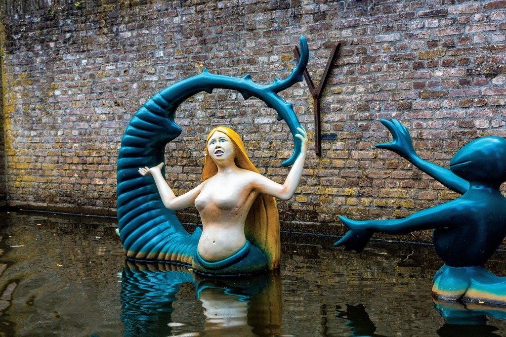 An outdoor, water-level view of public sculptures positioned in a calm canal against a tall, weathered brick wall. The central sculpture depicts a blonde mermaid-like creature with an open mouth, emerging from a curved, ribbed blue-green tail structure. To her right, another dark blue, frog-like creature stands in the water with outstretched, webbed hands reaching toward her. The dark canal water creates clear reflections of both whimsical sculptures.