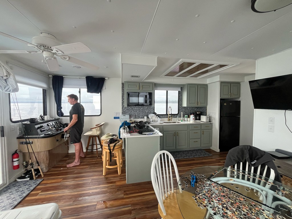A wide-angle interior shot of a spacious, modern houseboat cabin with a man piloting from the lower helm console on the left. The open-concept living space features warm wood-look flooring, white ceilings with a ceiling fan, and a full kitchen in the center with light sage green cabinets, a kitchen island with barstools, a black refrigerator, and a microwave. In the foreground, a glass-top dining table holds an unfinished jigsaw puzzle, while a flat-screen television is mounted on the wall to the right. Windows throughout the cabin reveal views of the surrounding open water under an overcast sky.