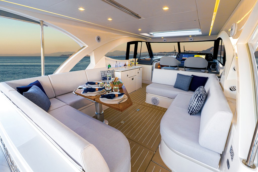 A bright, wide interior view of the main deck salon and cockpit lounge area on a modern yacht. The space is lined with light-tan teak-style flooring and features two large, plush, U-shaped sofas upholstered in light grey fabric, accented by navy and patterned blue throw pillows. On the left, a folding wooden dining table is neatly set with dinnerware and rolled blue napkins. In the background, a small wet bar console with drinks stands near the forward helm station, where two grey captain's chairs face the windshield. Large open side windows offer a panoramic view of a calm blue ocean during a sunset.