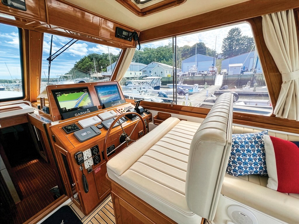 A bright, wide-angle interior view of the helm station and salon inside a classic motor yacht. The space is heavily defined by high-gloss, polished teak wood paneling and cabinetry. On the left, the helm console features twin Garmin digital chartplotters and navigation screens, electronic throttles, and a traditional stainless steel destroyer steering wheel. A wide, cream-colored leather bench seat with ribbed stitching serves as the helm seating, continuing into a salon sofa on the right adorned with blue-and-white patterned and solid red accent pillows. Large wrap-around windows offer a clear view of a busy marina dock with boats and shipyard buildings under a partly cloudy sky.