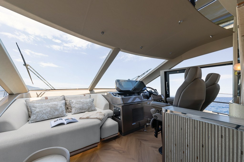 An interior view of the main deck helm station and forward salon seating area inside a luxury Galeon 620 Fly motor yacht. To the left, an open magazine rests on a large, cream-colored L-shaped companion sofa decorated with patterned accent pillows under massive, angled front windshield windows that look out to a bright sky. On the right, the main helm console features a dark dashboard panel with digital multi-function displays, a steering wheel, and dual premium tan leather captain's chairs. The cabin is finished with a modern upholstered ceiling panel and warm, herringbone-patterned wood flooring, with a side exit door visible next to the captain's station.