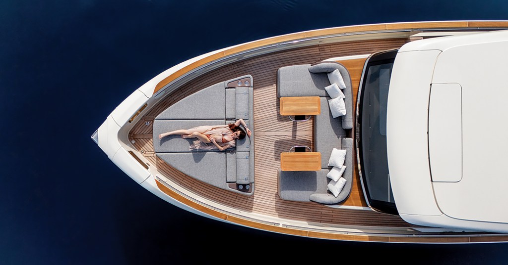 A top-down, aerial drone shot of the luxurious teak-planked foredeck of an Invictus ST550 yacht floating on deep blue water. A woman in a swimsuit and cover-up lies sunbathing on a massive, triangular grey sunpad built into the bow section. Just behind her, a spacious U-shaped grey lounge seating area features white accent pillows and two small, polished wooden dining tables facing the glass windshield of the enclosed white cabin.