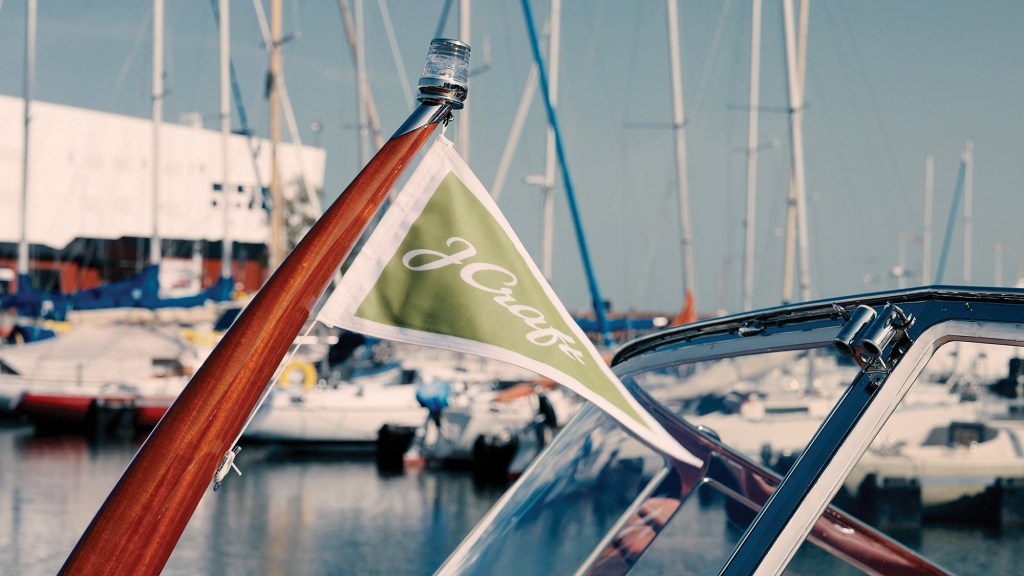 A close-up, shallow-depth-of-field shot focusing on a triangular green burgee flag featuring script text that reads "J Craft." The flag is attached to a polished wooden staff with a chrome navigation light at the tip, positioned just above the boat's chrome-framed glass windshield. In the blurred background, numerous sailboats with tall masts are moored in a sunny marina.