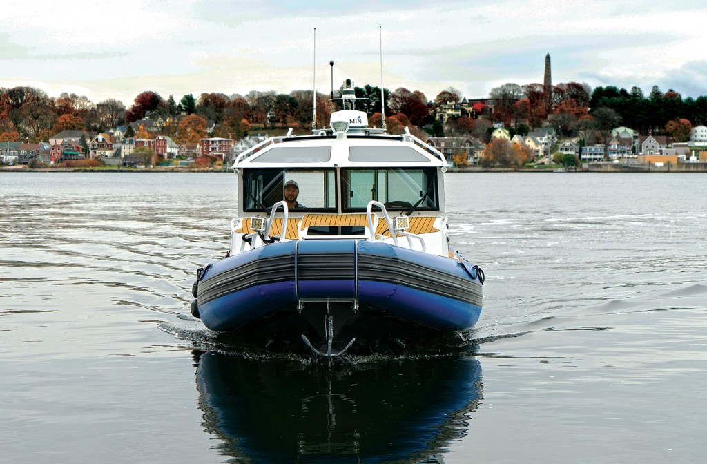 A head-on, eye-level shot of a white Life Proof rigid inflatable boat (RIB) cruising slowly forward on calm water, casting a clear reflection of its blue and black inflatable bow collar. The vessel features a faux-teak forward deck, bow rails with mounted LED spot-lights, and an enclosed cabin where the captain is visible behind the windshield. The cabin roof is equipped with a Garmin radar dome and dual antennas. In the background across the water, coastal houses sit nestled among autumn-colored trees on a hillside featuring a tall stone monument obelisk.