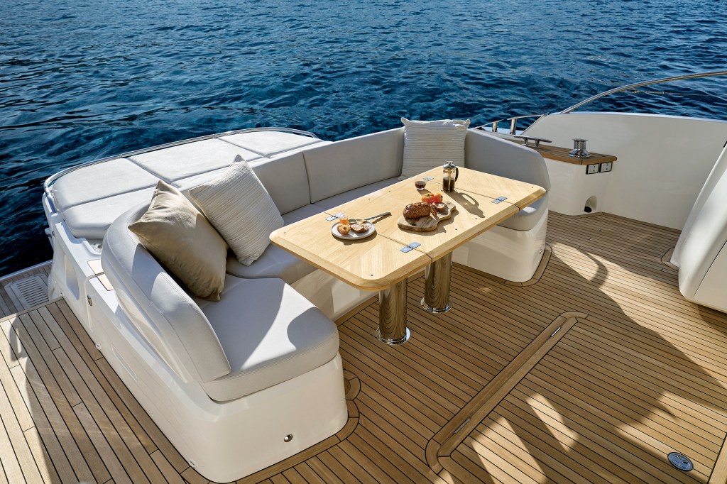 Spacious aft cockpit of a Princess V65 yacht featuring a U-shaped upholstered dining sofa, an adjustable teak wood table set with breakfast food, and a large sunpad leading to the open blue sea.