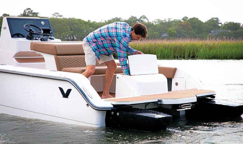 A close-up shot of the aft deck on a white Volare catamaran idling in shallow water. A man in a pink and blue plaid shirt and shorts leans over to open a small, white rectangular storage compartment lid built into the transom wall. Behind him, a rear-facing tan bench seat sits adjacent to the white helm console and black steering wheel. The stern features a light brown faux-teak swim platform extension sitting directly above two black hull pods, set against a background of green marsh grass.