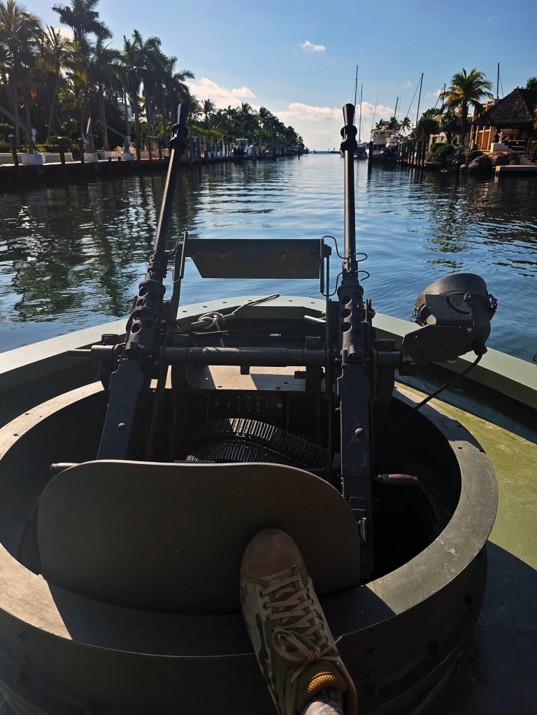 A first-person, point-of-view shot from the gunner's turret at the bow of a military patrol boat cruising down a calm tropical canal. Prominently featured in the foreground is a dual .50-caliber machine gun mount aimed forward, with a single camo-patterned shoe resting on the turret armor plate. The boat moves smoothly between rows of waterfront docks lined with tall palm trees and luxury homes. The clear water reflects the bright blue sky and distant clouds under bright, direct sunlight.