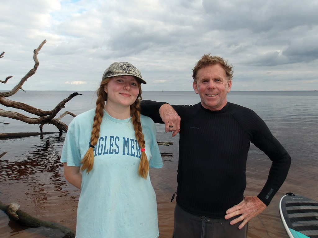 A medium, eye-level outdoor shot of a young woman and an older man standing close together on a shoreline under a cloudy sky, smiling for a photo. The woman, on the left, wears a light blue t-shirt with text ending in "...GLES MER", a camouflage baseball cap, and has her blonde hair styled in two long braids. The man, on the right, leans his arm casually on her shoulder while wearing a black long-sleeve rash guard or wetsuit top and dark shorts. In the background, a large piece of gnarled driftwood rests in the shallow, tea-colored water, which stretches out to a flat horizon, with the edge of a paddleboard visible on the bottom right.