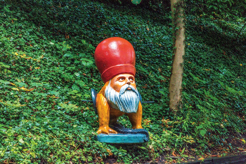 A whimsical outdoor sculpture of a creature nestled against a steep, heavily green hillside. The colorful sculpture features a human-like head with a white beard, a mustache, and a very large, rounded red hat or crown. This head is set onto a yellowish-brown quadrupedal animal body with clawed feet, standing on a blue base. A thin tree trunk stands to the right, and the entire background is covered in a dense carpet of green climbing ivy and foliage.