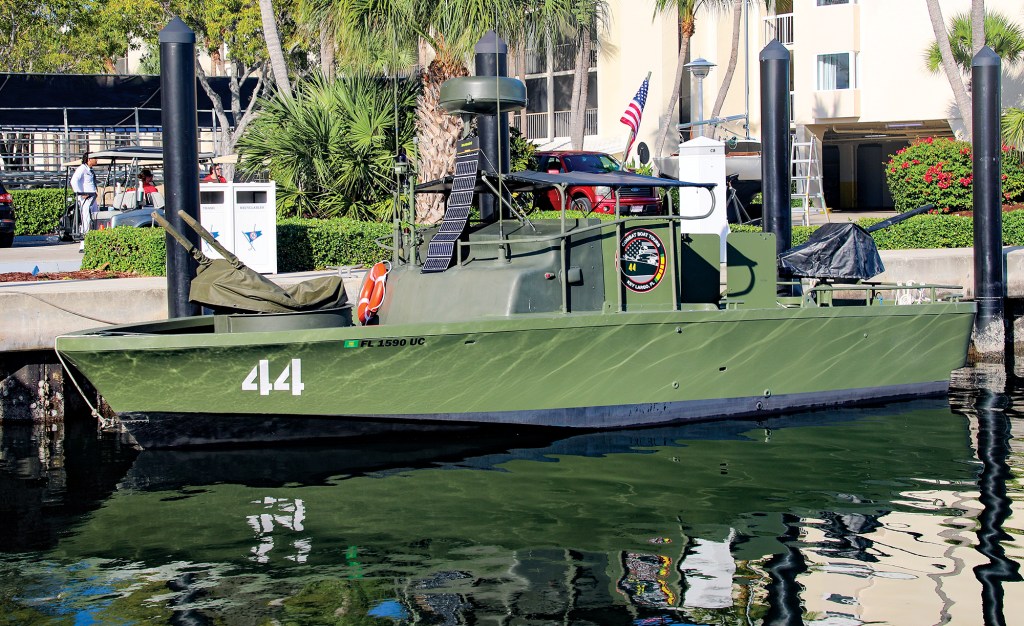 A side-profile view of an olive drab green military patrol boat docked in a marina alongside a concrete seawall. The vessel features the white hull number "44" stenciled near its bow and a black lower hull line. It is equipped with covered weapon mounts on the bow and stern, an orange life ring, a circular radar dome, and a flexible solar panel strip mounted vertically against the helm structure. An American flag flies from a staff at the stern. In the background, palm trees, a red vehicle, a golf cart, and a white building frame the sunny dock scene, with a clear reflection of the boat dancing on the water's surface.