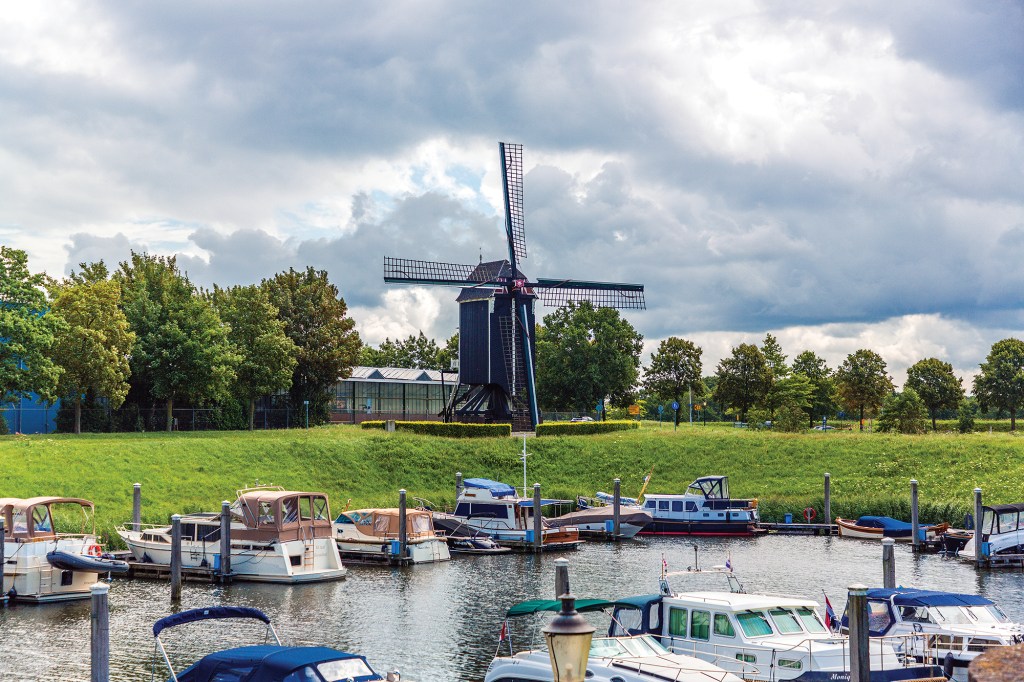 An outdoor, slightly elevated shot of a small marina filled with various motorboats and cabin cruisers docked along low piers. In the background, a steep green grassy bank rises up to a line of trees, behind which stands a historic black wooden windmill with its sails fully extended. The entire scene is set under a dramatic, overcast sky filled with dense gray and white clouds.