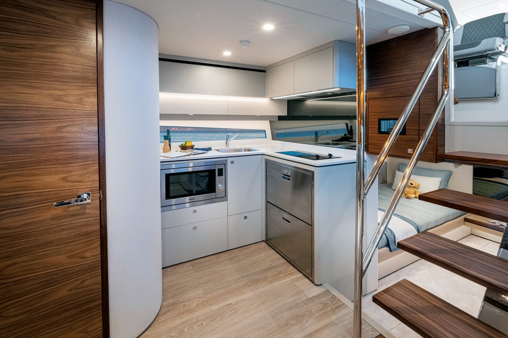 An interior view of a modern, compact lower-deck galley kitchen aboard a yacht. The space features light-colored wood flooring, minimalist white cabinetry, a white countertop with an integrated cooktop, a small sink, a built-in stainless steel microwave, and double drawer-style refrigerators. On the left is a curved wall leading to a rich grain wood door, while a modern set of open wooden floating stairs with stainless steel railings stands on the right. In the background behind the stairs, a cozy guest cabin berth is visible, neatly made with light blue bedding.