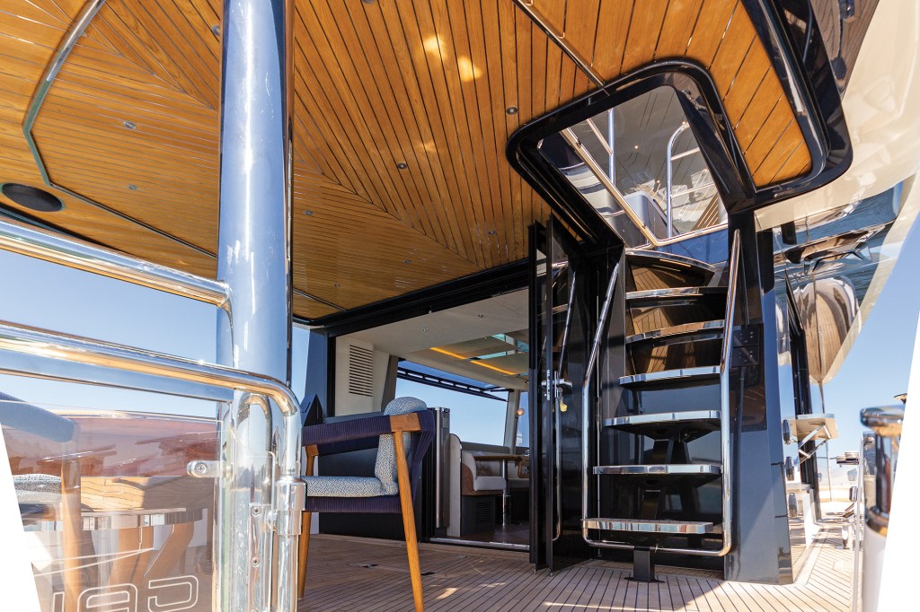 A low-angle, upward view looking from the teak aft deck cockpit toward the flybridge access staircase on a luxury Galeon 620 Fly motor yacht. On the right, a contemporary floating spiral staircase features black structural supports and wide, polished stainless steel open steps leading up through a square ceiling hatch. The expansive cockpit overhead is lined with glossy, varnished wood planks laid out in a chevron pattern and embedded with small LED downlights. Polished chrome structural pillars and safety handrails border the teak deck, which opens up through dark glass doors into the main salon cabin.
