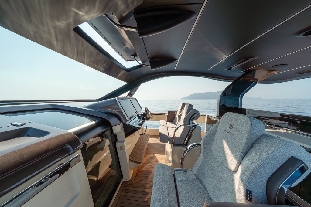 An interior shot looking forward through the modern, enclosed bridge helm station of an Invictus ST550 yacht. In the foreground, a premium light grey textured helm chair with an embroidered Invictus logo sits to starboard, while a matching double passenger bench is positioned to port. The sleek helm console is equipped with two large black glass multi-function displays, steering wheel, and throttle controls. Teak-planked flooring runs underfoot beneath a sculpted, dark-paneled ceiling, and large wrap-around glass windshields look out onto a calm, sunny blue ocean with a distant island hill.