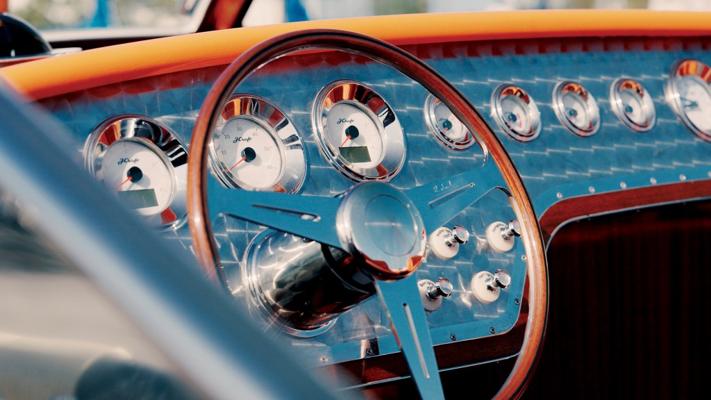 A close-up view of a classic-styled boat dashboard featuring an engine turned, swirled metallic helm panel. A polished wooden steering wheel with chrome spokes sits in front of circular, chrome-rimmed gauges with white faces labeled "J Craft." Retro-style chrome toggle switches are mounted to the right of the steering column, beneath a bright orange upholstered dash trim.