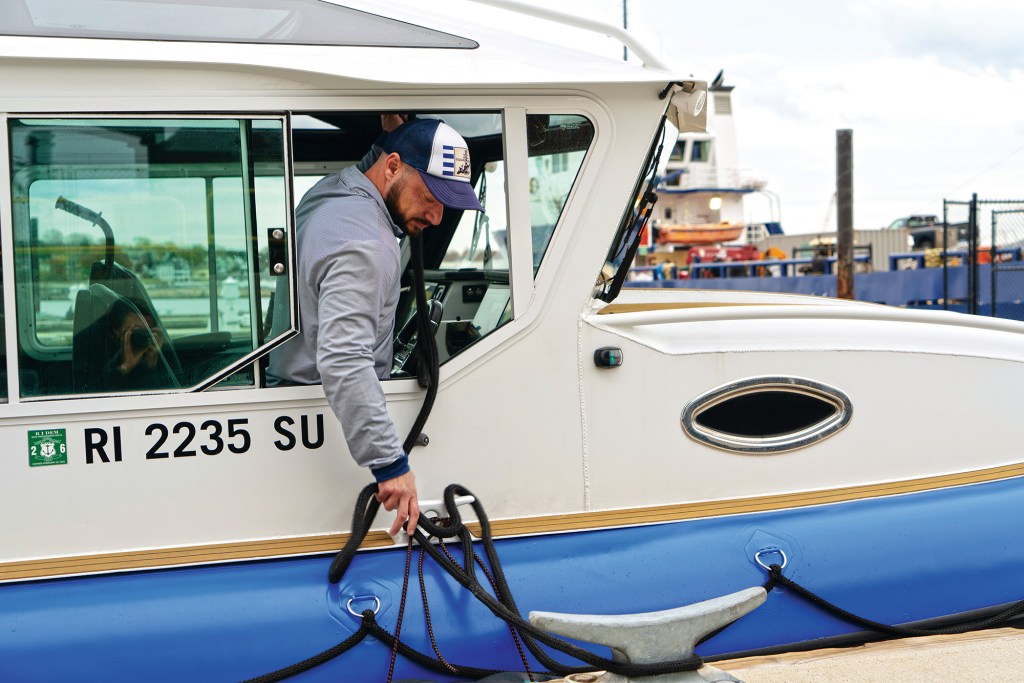 A close-up side view of a man in a blue and white baseball cap and a striped long-sleeve shirt leaning out of the open window of a white Life Proof cabin cruiser. He is looking down to secure a heavy black dock line to a deck cleat on the boat's gunwale, situated right above the bright blue inflatable collar hull. The boat's registration number, "RI 2235 SU," is visible on the cabin side, and a large metal dock cleat rests in the foreground on the right.