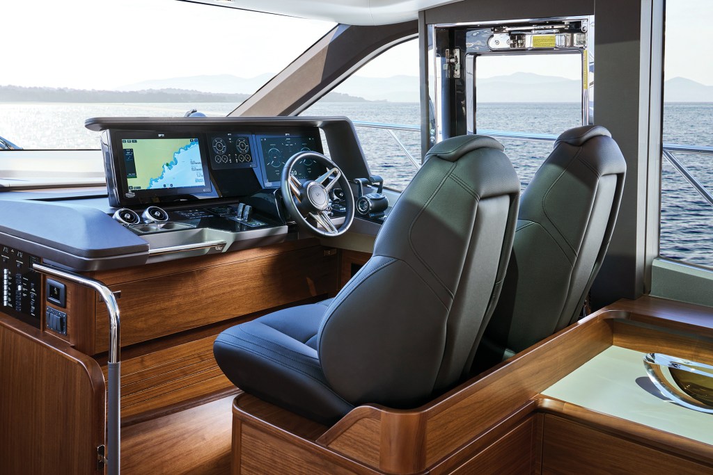 Modern helm station of a Princess V65 yacht featuring dual black leather captain's chairs, a steering wheel, multi-screen electronic navigation displays showing digital charts, and large windows with an open sea view.