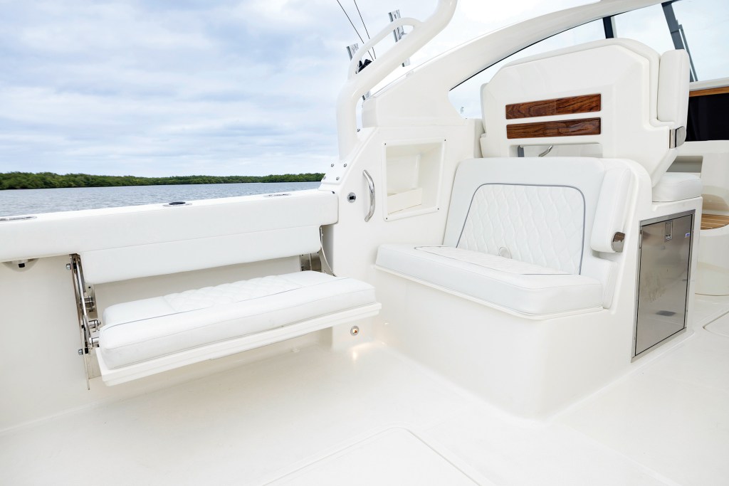A medium shot showcasing the white cockpit and transformational seating arrangement on a Pursuit DC 286 boat. To the left, a plush, white quilted gunwale bench is extended into its open seating position on stainless steel hinges. To the right, an integrated rear-facing seat with matching diamond-quilted cushioning is built into the helm structure, featuring wood trim accents on its headrest and a stainless steel storage door beneath the side seat. In the background, calm water and a green shoreline are visible under a cloudy sky.