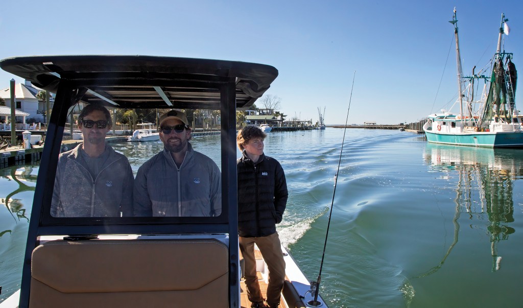 A medium shot from behind the helm seat looking forward as three men cruise down a sunny canal channel on a Volare boat. Two men wearing sunglasses and grey fleece jackets stand side-by-side behind the glass windshield of the center console helm under a dark T-top canopy. A third man in a black puffer jacket looks off to the right from the deck walkway. A single fishing rod is mounted in a gunwale holder, and a large, green-and-white commercial shrimping boat is docked on the right side of the calm water channel under a clear blue sky.