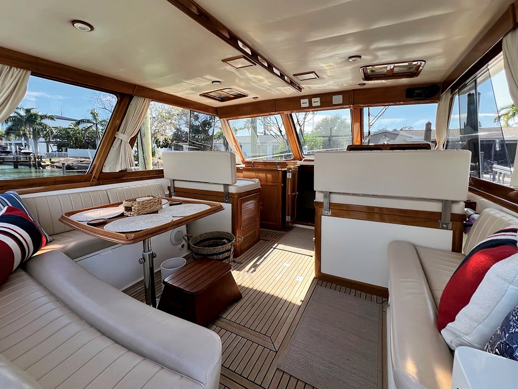An interior view looking forward through the sunlit salon of a classic yacht toward the helm. The space features traditional teak-and-holly flooring, high-gloss wood cabinetry, and large panoramic side windows draped with white curtains. On the left, a U-shaped cream leather dinette sofa surrounds a polished wooden pedestal table set with woven placemats and a small basket. On the right, a matching straight sofa sits across the aisle. Further forward, two wide, cream-upholstered bench seats face the front windshield windows, which reveal a bright, tree-lined tropical coastline outdoors.