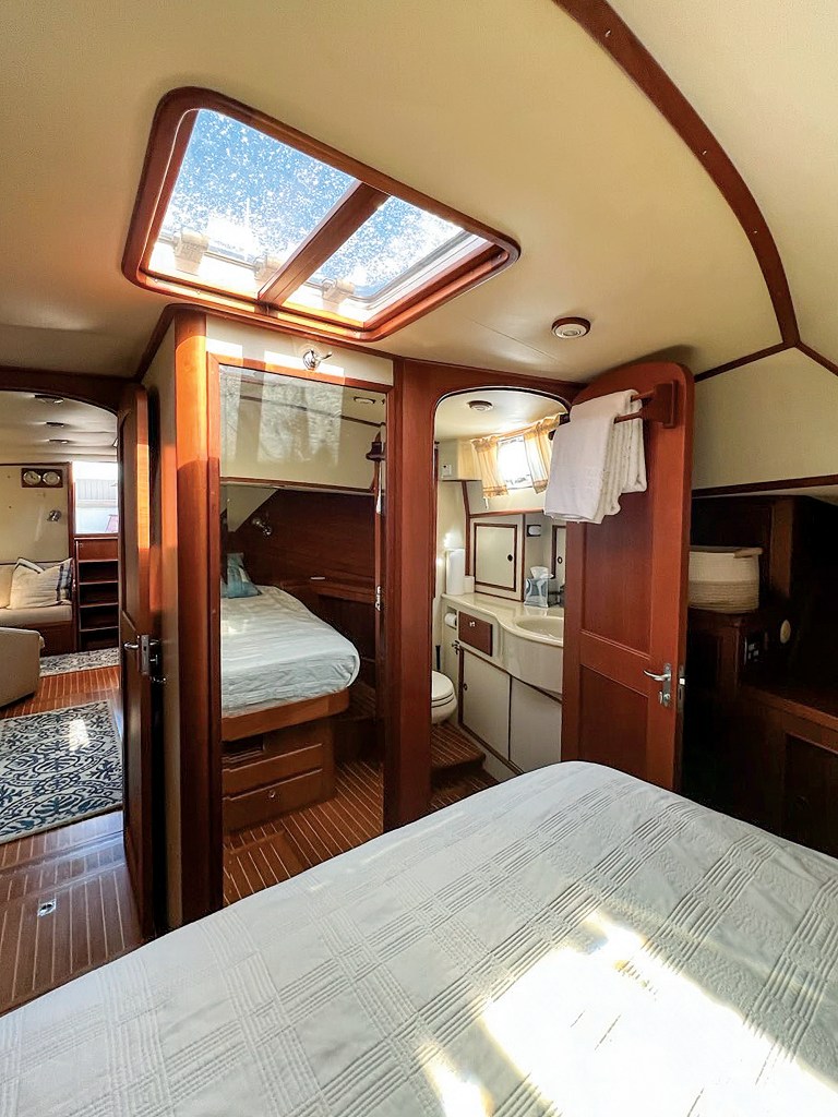 An interior view looking out from the master stateroom into the lower cabin accommodations of a classic yacht. The master bed with a quilted white coverlet rests in the foreground. The hallway features traditional teak-and-holly flooring and warm, satin-finished wood paneling under a cream-colored arched ceiling with a large wooden-framed skylight hatch. Open doorways reveal a guest cabin with a single berth to the left and a compact head compartment with a vanity sink and toilet to the right. In the far background, a lower lounge area with patterned area rugs and a sofa is visible.