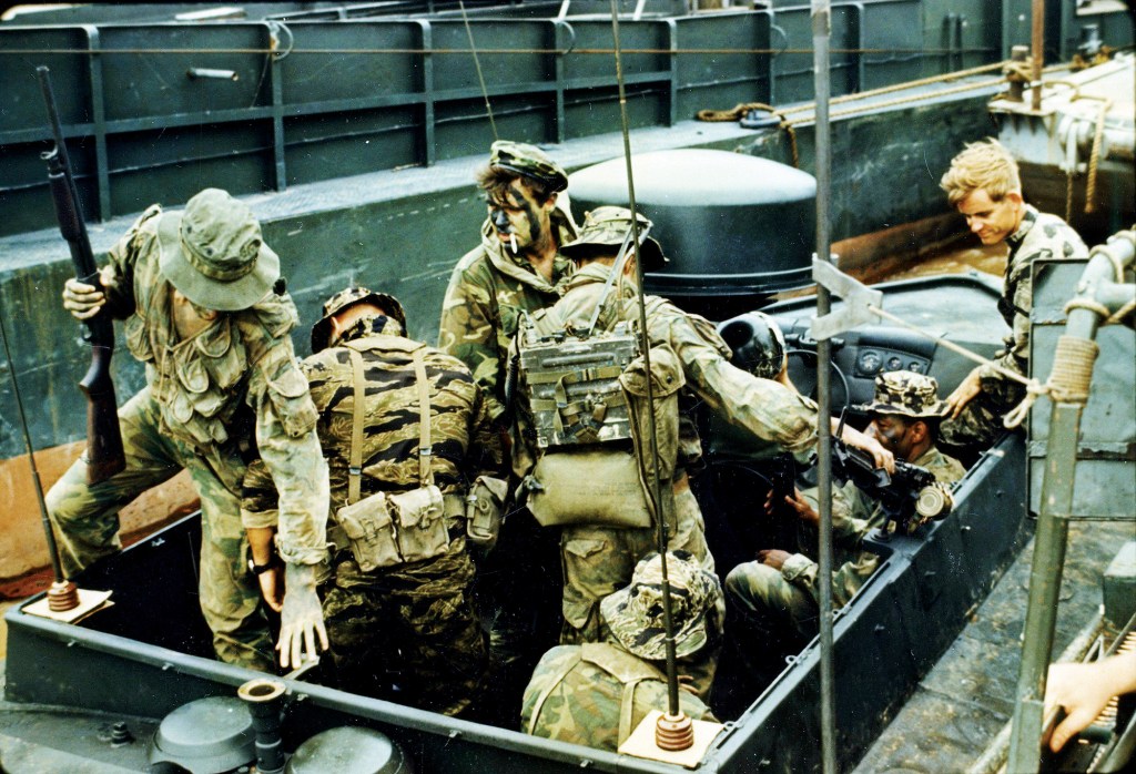 A historic, candid photograph from the Vietnam War era showing a group of American soldiers crowded on a small combat patrol boat. The soldiers wear camo jungle fatigues, tiger-stripe patterns, boonie hats, and tactical gear, with several men sporting dark camouflage face paint. One soldier on the left steps onto the boat while holding a shotgun aloft, another in the center carries a heavy military radio pack on his back, and others are stationed around the helm and weapons areas. A large black radar dome sits prominently behind the helm, all framed against a dark metal vessel hull in the background.