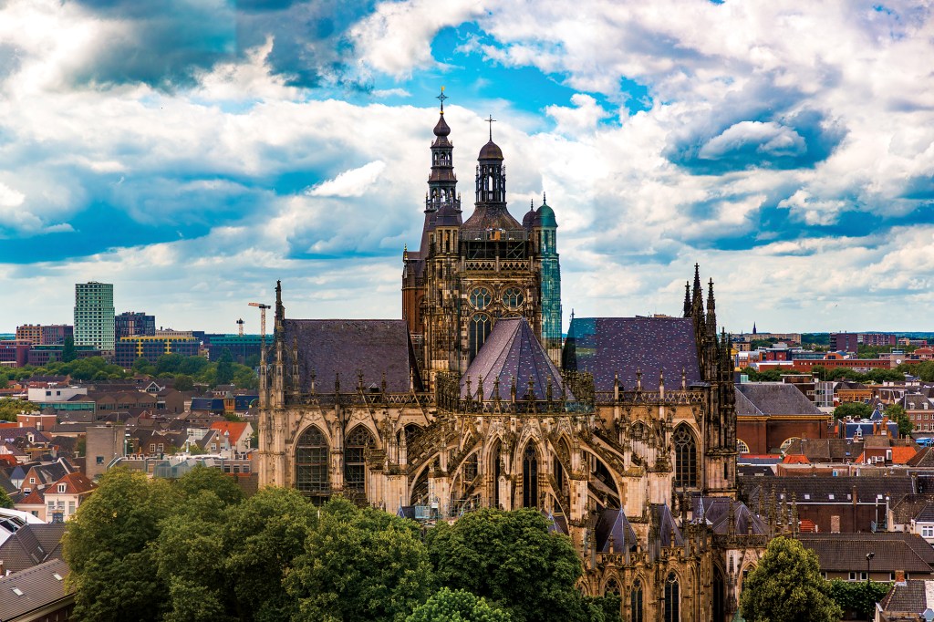 A breathtaking, high-angle panoramic view dominating a massive historic Gothic cathedral surrounded by a sprawling European city. The cathedral features intricate stone carvings, pointed arches, large stained-glass windows, soaring spires, and a multi-tiered central tower topped with crosses. The lower portion of the cathedral is framed by lush, green treetop canopies. In the background, a dense mix of traditional brown-roofed houses and modern high-rise city buildings stretches to the horizon under a dramatic, bright blue sky filled with puffy white and gray clouds.