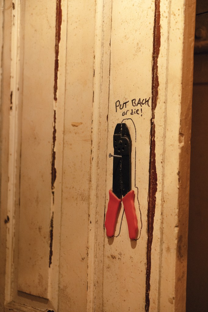 A pair of wire strippers with red handles hanging from a screw on a weathered white wooden door, with an outline drawn around the tool and handwritten text reading "PUT BACK or die!".