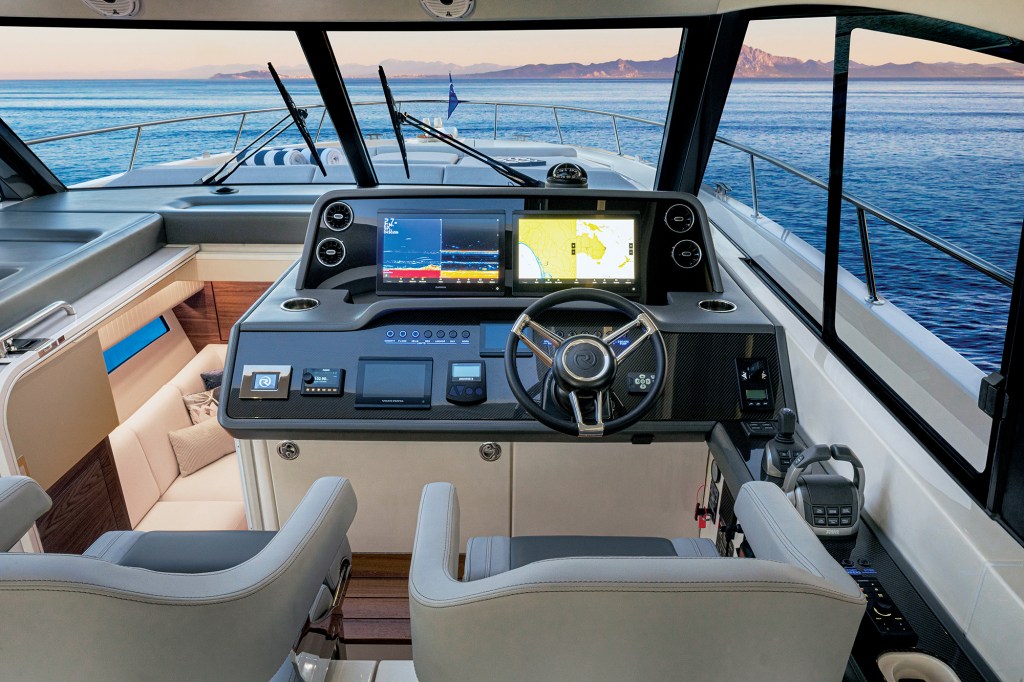 A high-angle, first-person view of a modern, high-tech yacht helm station. The matte-grey dashboard features a carbon-fiber accent panel, a black leather-trimmed steering wheel with a silver center logo, electronic engine throttles, and a joystick control. Two large multifunction display (MFD) screens are illuminated, displaying sonar and navigation chart data. To the left of the helm console, a companionway opens up to show a glimpse of a lower lounge cabin with a cream-colored sofa. In the foreground, the backs of two light-grey leather captain's chairs are visible, while the expansive forward windshield looks out over the bow toward a calm, vast blue ocean with mountains on the distant horizon during dusk.
