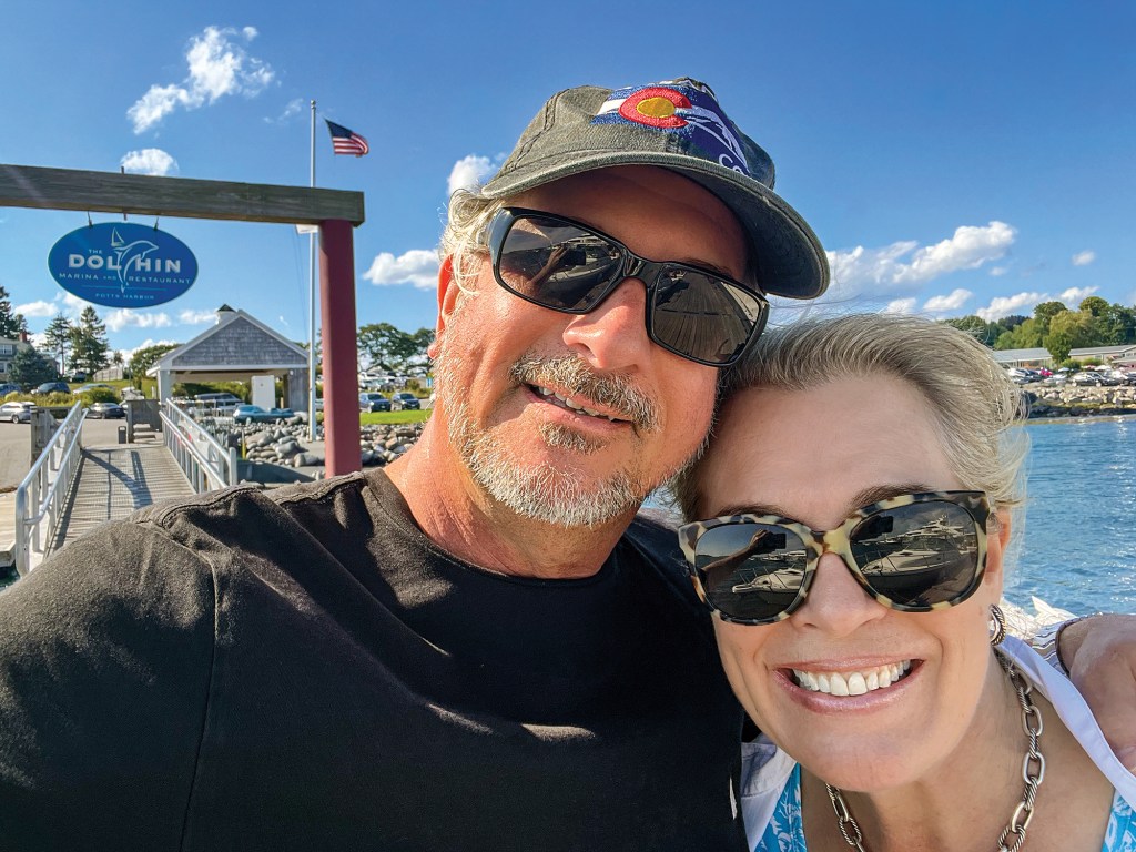 A sunny, close-up selfie of Steven and Leah Kovich smiling together outdoors near a waterfront. On the left, Steven wears a black t-shirt, dark sunglasses, and a grey baseball cap featuring a colorful Colorado logo patch. Leah, leaning close to him on the right, smiles broadly while wearing tortoiseshell sunglasses, a silver chain necklace, and a blue-and-white patterned top. In the background, a wooden dock ramp leads to a sign that reads "The Dolphin Marina and Restaurant," set against a bright blue sky with fluffy white clouds, green trees, and calm coastal water.