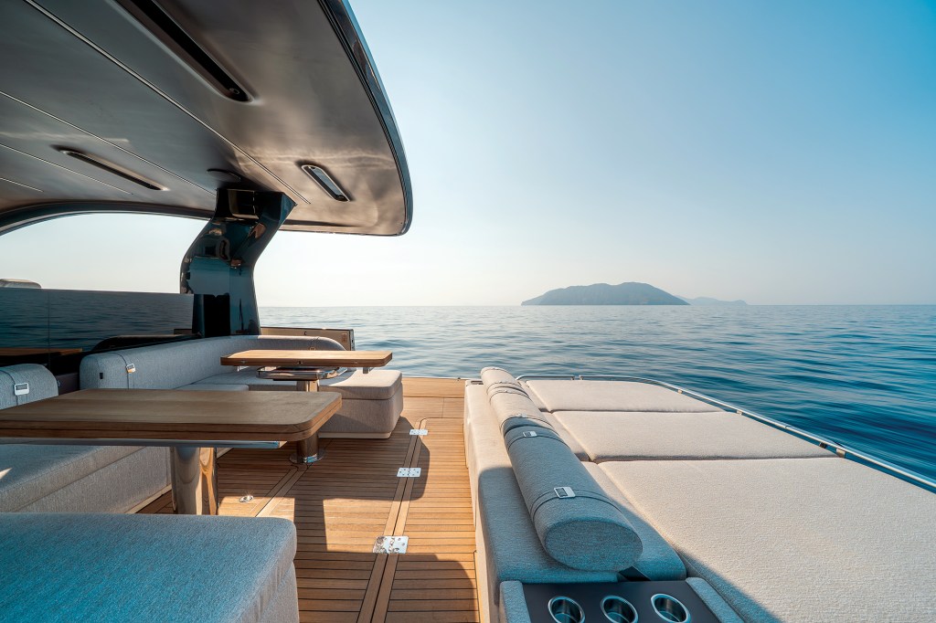 A wide shot looking aft onto the open teak-planked cockpit and stern deck of an Invictus ST550 yacht. On the left, a grey upholstered L-shaped dining lounge features two rectangular wood-topped tables on polished stainless steel pedestals beneath a sleek hardtop canopy extension. To the right, a massive, flat sunpad lounger extends toward the stern, lined with cylindrical headrest bolsters and built-in cup holders. The open deck faces a calm, wide blue ocean under a hazy sky with a single hilly island on the horizon.