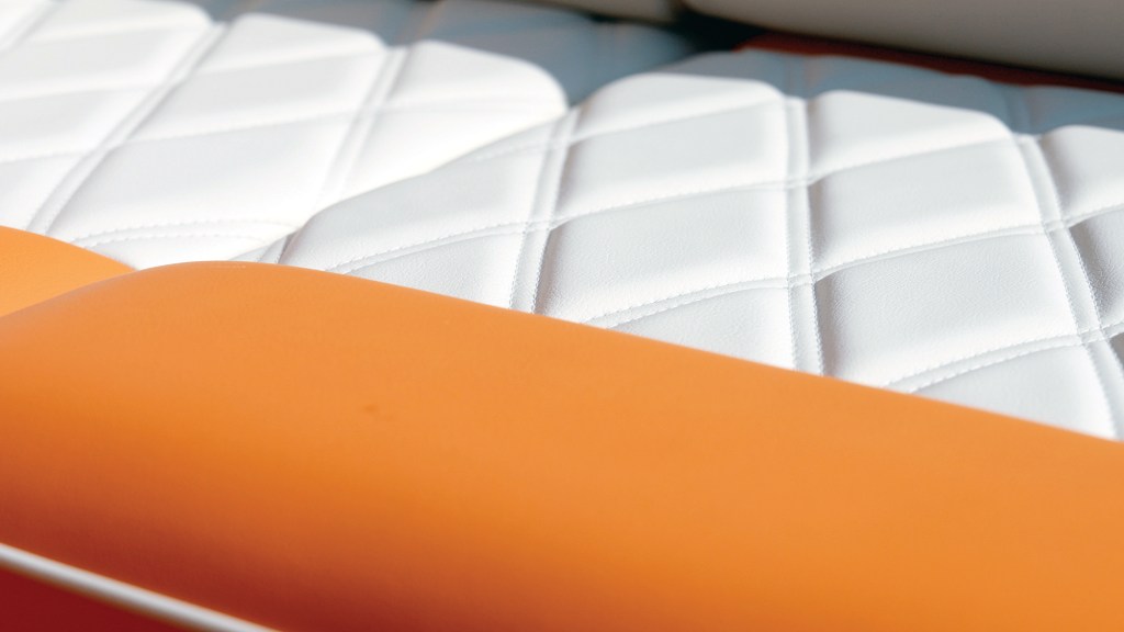 An extreme close-up, abstract shot of luxury boat upholstery. The foreground is dominated by a smooth, vibrant orange leather bolster cushion. Behind it, the seating features premium white leather stitched in a detailed diamond-quilted pattern, with a soft-focus hint of grey or blue paneling visible at the very top edge.