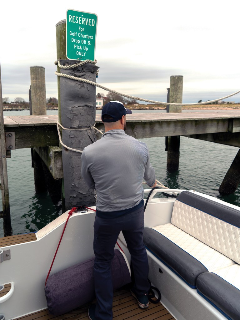 A full-length, high-angle shot from behind a man standing on the stern deck of a white boat as it pulls up alongside a wooden dock piling. The man wears a navy baseball cap, a grey long-sleeve shirt, and dark pants while handling a dock line. A green sign mounted on the weathered piling reads, "RESERVED For Golf Charters Drop Off & Pick Up ONLY." The boat's cockpit features diamond-stitched white and navy upholstery and a faux-teak floor, with a purple fender resting at the man's feet.