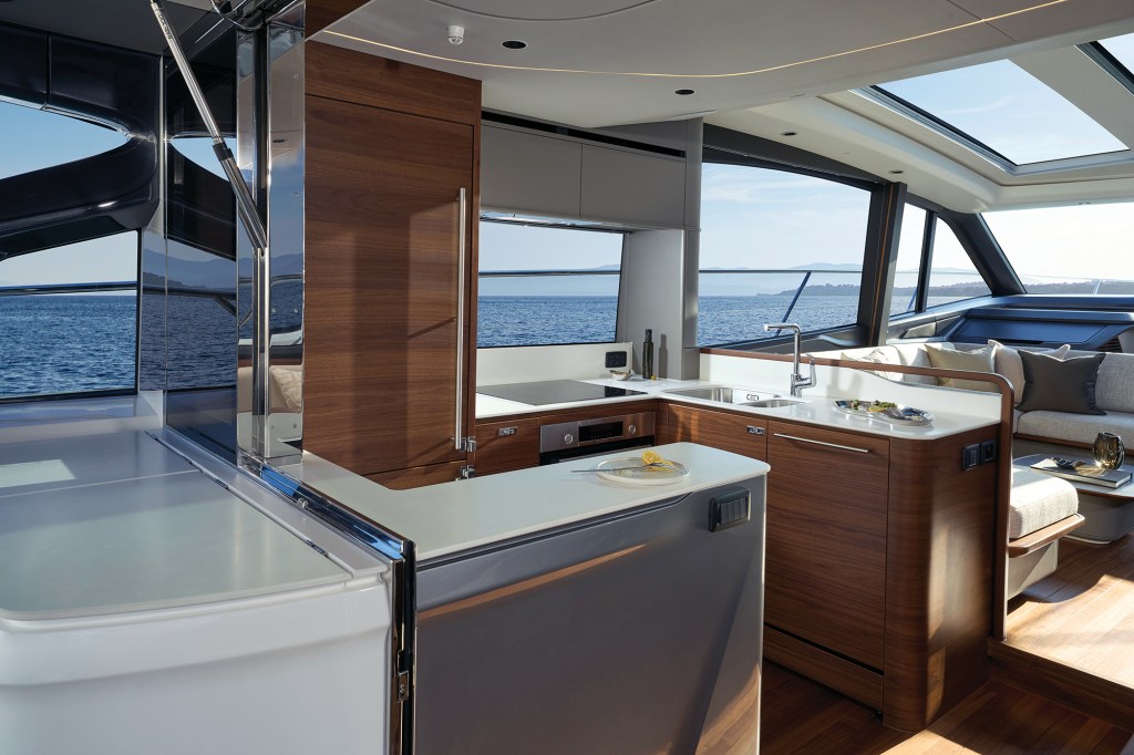 Contemporary aft galley on a Princess V65 yacht featuring clean white countertops, integrated stainless steel appliances, premium wood cabinetry, and an open layout that connects smoothly to the saloon under an overhead sunroof.