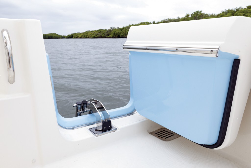 A close-up shot of the starboard-side dive and boarding door swung wide open on a Pursuit DC 286 boat. The heavy white hull door features a light blue fiberglass interior matching the exterior hull color, lined with a thick black rubber seal. A robust stainless steel hinge mechanism secures it to the deck, creating a seamless opening directly to the water. In the background, calm grey river water extends toward a lush green mangrove shoreline under an overcast sky.