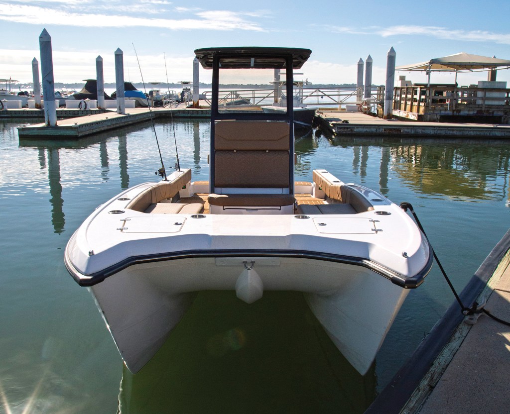 A head-on, low-angle shot of the distinct twin-hull bow of a white Volare center-console power catamaran tied to a concrete dock. The open bow deck features two integrated storage hatches and leads back to a central tan upholstered lounge seat positioned on the front of the helm station. A black T-top canopy covers the helm console, and two fishing rods are visible sitting in gunwale holders. In the background, calm marina water reflects vertical white pilings, floating docks, and a covered pavilion under a bright blue sky.